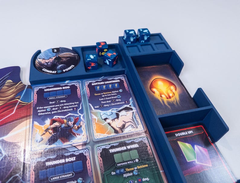 May include: A blue plastic board game organizer containing four six-sided dice with colored symbols, a circular combat points token, and several rectangular game cards featuring illustrations of a character with a hammer and lightning effects.