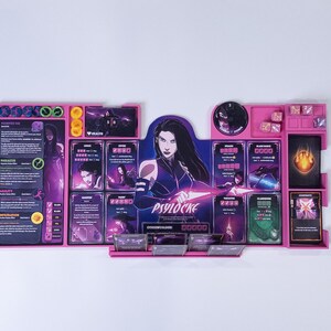 May include: A pink plastic board game organizer for Psylocke featuring dedicated slots for character cards, tokens, and dice. The center displays a large illustration of the character Psylocke with purple and black accents, alongside various game components and text labels.