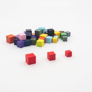 May include: A collection of small, solid-colored wooden cubes in various hues including red, blue, yellow, green, purple, pink, orange, black, and grey. The cubes are arranged in a loose pile with three individual red cubes placed in the foreground.