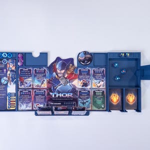 May include: A blue plastic board game organizer featuring a central character illustration of Thor with a hammer. The tray includes designated slots for game cards, small circular tokens, and dice, with printed text detailing character abilities.