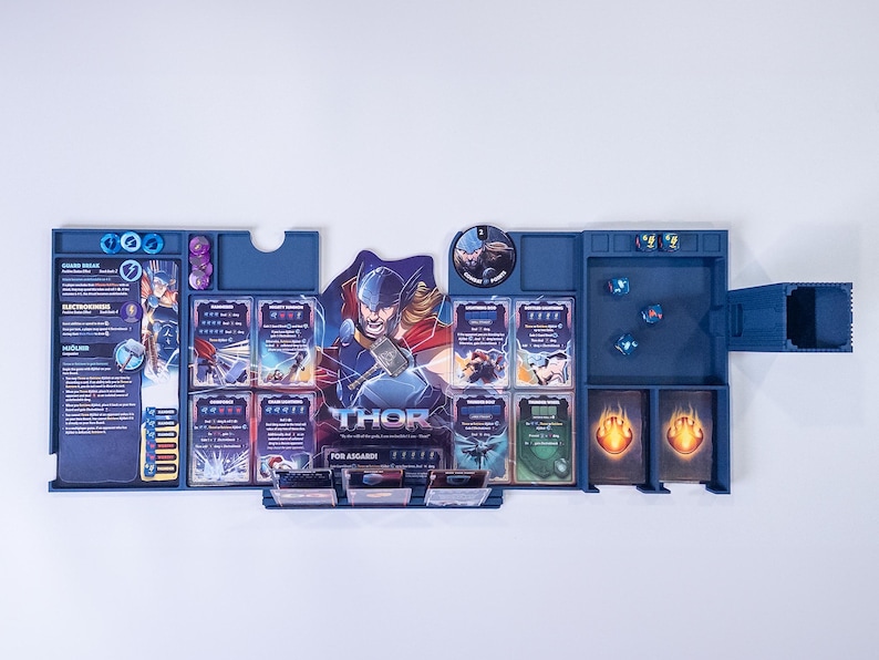 May include: A blue plastic board game organizer featuring a central character illustration of Thor with a hammer. The tray includes designated slots for game cards, small circular tokens, and dice, with printed text detailing character abilities.