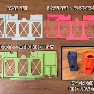 May include: A light blue base set, a pink base set with a card tray, a light green base set with a card and dice tray, and a dark blue and orange magnetic dice tower. The text BASE SET, BASE SET + CARD TRAY, BASE SET + CARD/DICE TRAY, and MAGNETIC DICE TOWERS are visible.