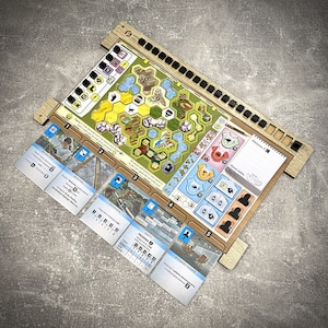 Può includere: Un tabellone da gioco in legno con una finitura marrone chiaro e un inserto per tabellone da gioco blu chiaro e bianco. Il tabellone da gioco ha un motivo a griglia con i numeri da 1 a 5. L'inserto del tabellone da gioco ha una mappa con una varietà di simboli e testo. Il tabellone da gioco ha anche un piccolo blocco note bianco con una penna nera.