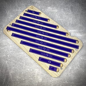 May include: A wooden game board with blue rulers marked from 2.5 cm to 28 cm. Each ruler has the text "GAMING FORGE" printed in white. The board has a logo in the top left corner and is set on a grey surface.