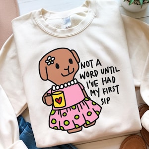 Könnte beinhalten: Ein weißer Pullover mit einer Cartoon-Illustration eines braunen Hundes, der ein rosa Kleid trägt und eine gelbe Tasse hält. Der Text "Not a word until I've had my first sip" ist auf dem Pullover aufgedruckt.