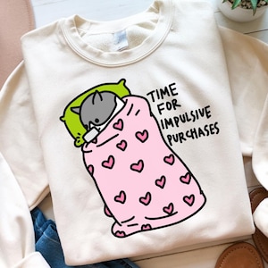 May include: A white sweatshirt with a cartoon illustration of a cat under a pink blanket with hearts. The text "TIME FOR IMPULSIVE PURCHASES" is printed on the sweatshirt.