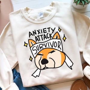 Op de afbeelding: Crèmekleurige sweatshirt met de tekst "ANXIETY ATTACK SURVIVOR" boven een cartoon hondenillustratie. De hond is oranje en wit met een verband op zijn hoofd. Het ontwerp bevat gele diamantaccenten.