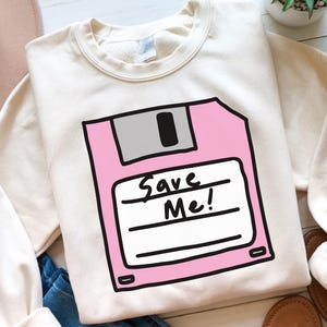 May include: A white sweatshirt with a pink floppy disk graphic. The floppy disk has a white label with the text "Save Me!" written on it.