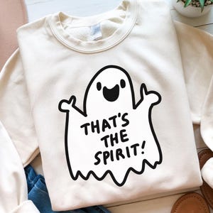 May include: Cream-colored sweatshirt with a black graphic of a smiling ghost and the phrase "THAT'S THE SPIRIT!" in black text. The sweatshirt has a crew neck and long sleeves, perfect for Halloween or spooky season.