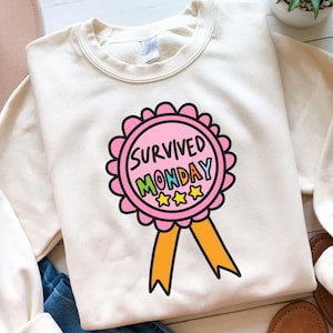 以下が含まれることがあります： 「SURVIVED MONDAY」という文字と3つの星が描かれたリボンがピンクとイエローでデザインされた白いスウェットシャツです。