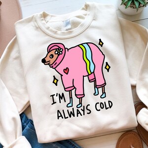 May include: Cream-colored sweatshirt featuring a cartoon dog in a pink outfit with a flower and heart design. The dog is wearing a pink hat and boots. The text "I'M ALWAYS COLD" is printed below the dog.