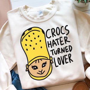 May include: A cream-coloured sweatshirt featuring a yellow Croc shoe design with a face, and the text "CROCS HATER TURNED LOVER" in black. This casual sweatshirt is suitable for everyday wear and is a comfortable style.