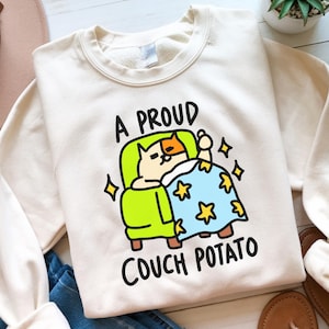 May include: Cream-colored sweatshirt with a graphic of a cat lounging on a green couch under a blue blanket with yellow stars. The text "A PROUD COUCH POTATO" is printed above and below the cat illustration. The sweatshirt is folded.