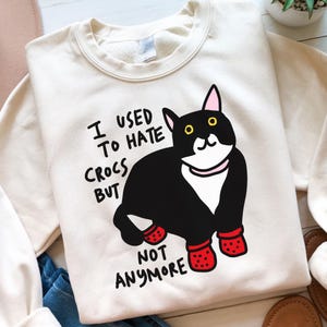 May include: Cream-coloured sweatshirt featuring a black and white cat wearing red Crocs. The text "I USED TO HATE CROCS BUT NOT ANYMORE" is printed on the front. The sweatshirt has a crew neck and long sleeves.