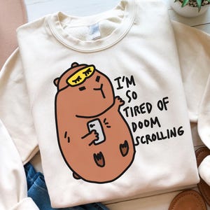 May include: A white sweatshirt with a brown bear wearing eye mask and holding a phone. The text "I'm so tired of doom scrolling" is printed on the sweatshirt.