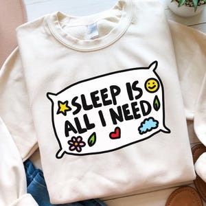 May include: A cream-colored sweatshirt with a graphic of a pillow that says "SLEEP IS ALL I NEED" with a star, flower, heart, leaf, and cloud design.