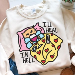 May include: A white sweatshirt featuring a cartoon illustration of a cat in bed with a glass of wine and a remote control. The text "I'll heal in hell" is printed on the sweatshirt.