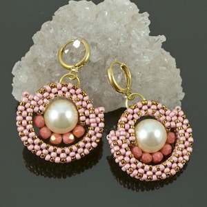 Puede incluir: Un par de pendientes Luna con un diseño de perlas y cuentas. Los pendientes presentan una gran perla blanquecina en el centro, rodeada de cuentas rosas y doradas. Los pendientes tienen aros dorados. El texto "Luna Earrings" y "BeadedTreasury" es visible.