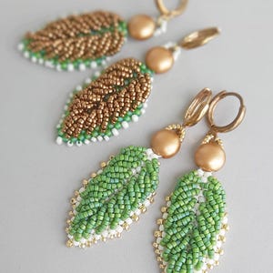 May include: Handmade beaded leaf earrings in gold and green. The earrings feature a leaf design with gold, green, and white beads. The earrings have gold-colored hoops and a gold bead accent. The text in the image reads "Dimensional Leaves beading pattern".