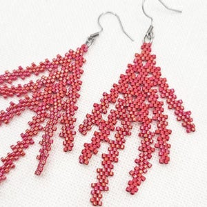 May include: A pair of coral-colored beaded earrings with a unique, branching design. The earrings are made of small, square beads and have silver-colored ear wires. The text "Playful Coral Earrings" and "BEADED TREASURY" is visible in the bottom right corner.