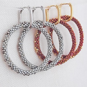 Puede incluir: Primer plano de tres pares de pendientes de aro con cuentas. Los pendientes son plateados, burdeos y dorados. Los pendientes plateados tienen cierres plateados, mientras que los pendientes burdeos y dorados tienen cierres dorados. Los pendientes cuelgan sobre un fondo blanco.