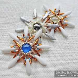 May include: Two handmade crystal snowflake brooches. Each brooch features a central jewel, surrounded by white, silver, and orange accents. The text "CRYSTAL SNOWFLAKE" and "©BEADED TREASURY 2022" are visible.