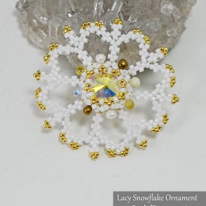 Könnte beinhalten: Ein filigranes Schneeflockenornament aus weißen Perlen mit goldenen Akzenten und einem zentralen irisierenden Kristall. Das Ornament hat ein zartes, offenes Design. Der Text auf dem Bild lautet "Lacy Snowflake Ornament @BeadedTreasury 2018."