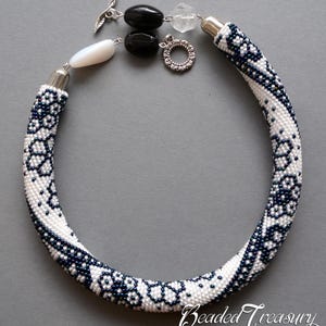 May include: A beaded necklace featuring a white and navy blue floral design. The necklace has a silver clasp and is decorated with black and clear beads. The text "Beaded Treasury" is visible in the bottom right corner.