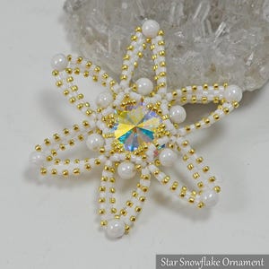 May include: A beaded star snowflake ornament with a central iridescent crystal. The ornament is white with gold and pearl accents. The text "Star Snowflake Ornament @BeadedTreasury 2018" is at the bottom.