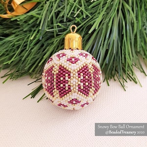 Könnte beinhalten: Ein mit Perlen besetzter Weihnachtsschmuck mit goldener Kappe und einem Muster aus weißen, bordeauxfarbenen und goldenen Perlen. Der Schmuck ist rund und liegt vor einem grünen Tannenzweig. Der Text auf dem Bild lautet "Snowy Bow Ball Ornament @BeadedTreasury 2020."