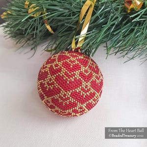 Op de afbeelding: Een rode, met kralen bezette kerstbal met een gouden geometrisch patroon, hangend aan een gouden lint en een groene dennentak. De bal is rond en heeft een textuur. De tekst "From The Heart Ball" en "BeadedTreasury 2022" is zichtbaar.