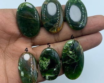 May include: Six polished, oval-shaped gemstones in shades of green and brown are displayed. The stones vary in pattern and color, with some featuring lighter green inclusions. The gemstones are held in a hand, showcasing their natural beauty.