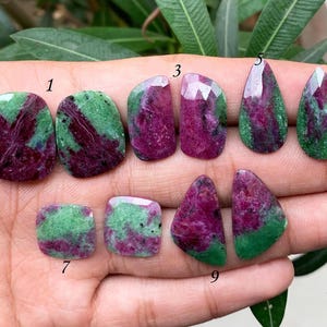 Pode incluir: Nove cabochões de pedras preciosas verdes e rosas de vários formatos e tamanhos, incluindo gota, quadrado e oval, dispostos em uma mão.
