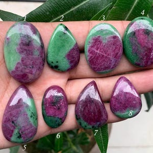 May include: Eight green and pink gemstone cabochons, some with a teardrop shape and others oval, are arranged on a hand. The cabochons are numbered 1 through 8.