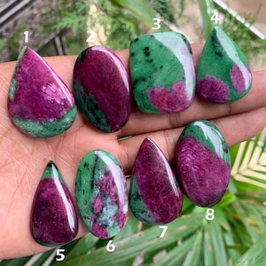 Könnte beinhalten: Eine Sammlung von acht polierten Zoisit-Edelsteinen mit Rubin-Einschlüssen. Die Steine haben unterschiedliche Formen, darunter Tropfen, Oval und Rechteck. Die Farben sind eine Mischung aus tiefem Magenta, Grün und Weiß.