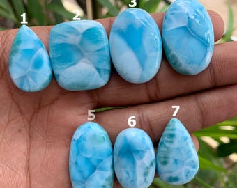 Può includere: Sette pietre preziose di larimar lucidate in varie forme, tra cui a goccia, quadrata e ovale. Le pietre mostrano una gamma di tonalità azzurre con venature bianche, che ricordano le onde dell'oceano. Le pietre preziose sono esposte su una superficie scura.