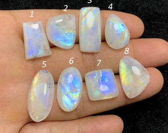 May include: Eight polished moonstone gemstones in various shapes, including rectangular, oval, and teardrop. The stones display a milky white base with iridescent blue and yellow flashes. The gemstones are held in a hand.