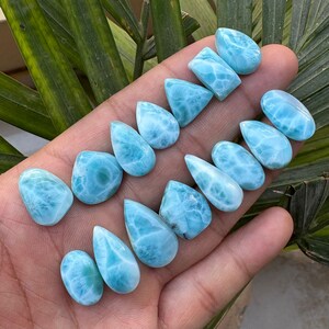 May include: A collection of polished, light blue larimar gemstones in various shapes, including teardrops, ovals, and rectangles. The stones display a marbled pattern of white and blue, with a smooth, glossy finish. The gemstones are displayed against a natural background.