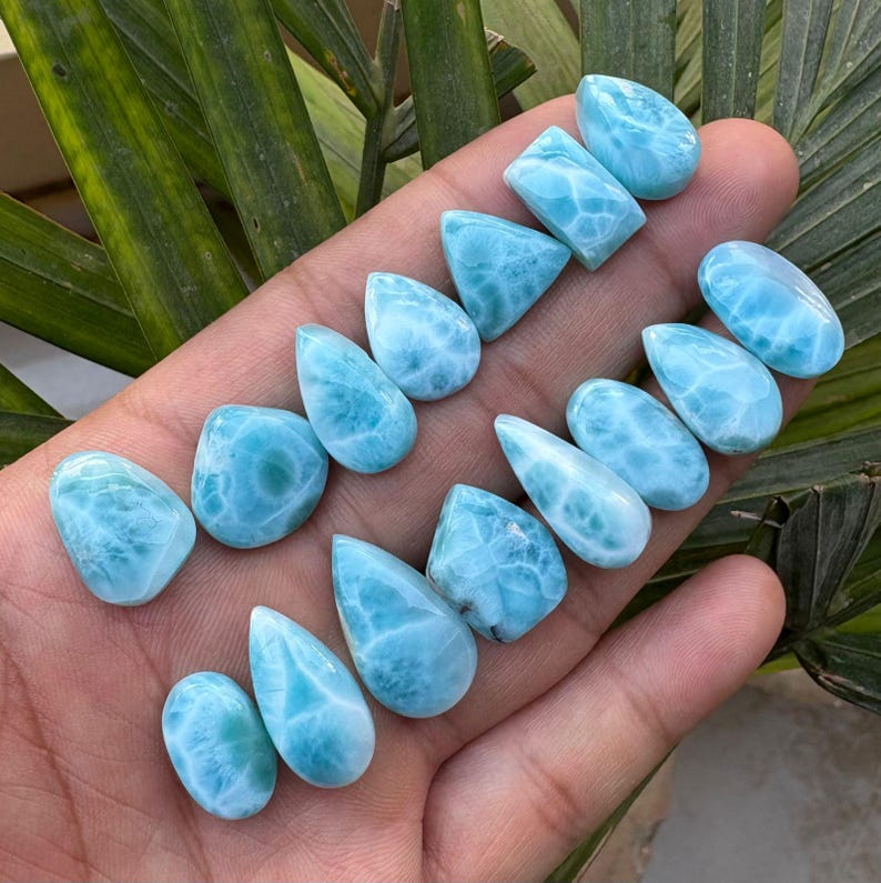 May include: A collection of polished, light blue larimar gemstones in various shapes, including teardrops, ovals, and rectangles. The stones display a marbled pattern of white and blue, with a smooth, glossy finish. The gemstones are displayed against a natural background.