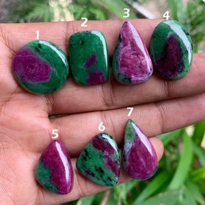 May include: A collection of seven polished ruby zoisite gemstones in various shapes. The stones feature a mix of deep green and vibrant magenta colors. The shapes include oval, rectangular, and teardrop designs, showcasing the natural patterns of the stones.
