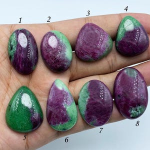 Puede incluir: Ocho cabujones de piedra preciosa verde y púrpura, numerados del 1 al 8, están dispuestos en una mano. Los cabujones son de forma ovalada y de lágrima.