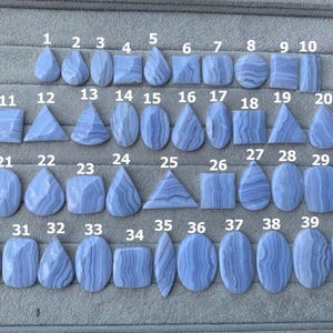 May include: Forty blue lace agate gemstones in diverse shapes, such as teardrops, squares, and ovals. Each stone features unique banding patterns and is numbered. The gemstones are displayed on a grey surface.