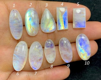 May include: Ten polished moonstone gemstones in various shapes, including oval, teardrop, and rectangular. The stones display a milky white base with iridescent flashes of blue, yellow, and green. The gemstones are numbered 1 through 10.