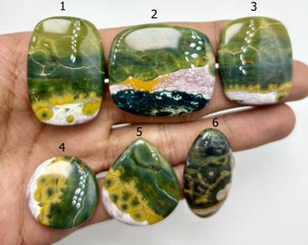 May include: Six polished gemstones in various shapes, including oval, rectangular, and teardrop. The stones feature a mix of green, yellow, and white colours with unique patterns. The gemstones are displayed against a neutral background.