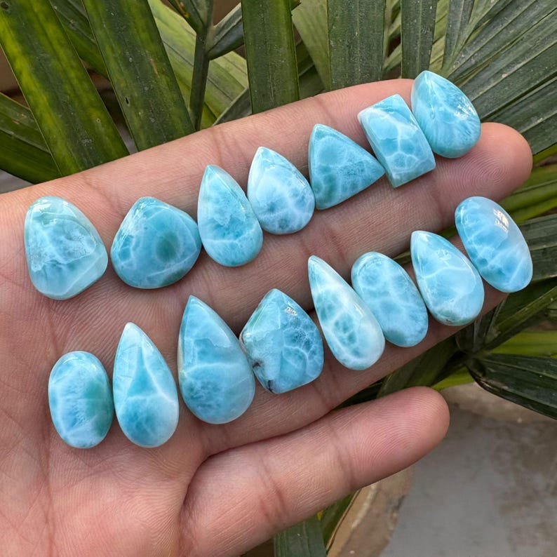 May include: A collection of polished larimar gemstones in various shapes, including teardrop, oval, and rectangular. The stones display a light blue color with white marbling, resembling ocean waves. The gemstones are displayed on a hand.