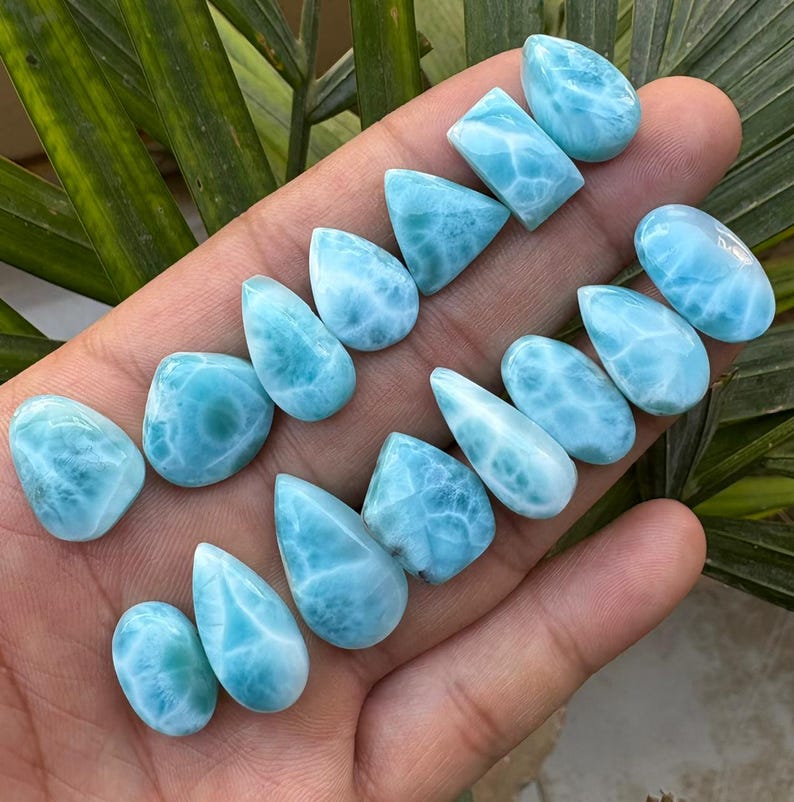 May include: A collection of polished larimar gemstones in various shapes, including teardrops, ovals, and rectangles. The stones display a light blue color with white marbling, resembling ocean waves. The gemstones are held in a hand against a backdrop of green leaves.