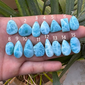 May include: A collection of fifteen polished larimar gemstones in various shapes, including teardrop, oval, and rectangular. The stones are a light blue color with white marbling, and are numbered 1 through 15.