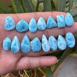 May include: A collection of polished larimar gemstones in various shapes, including teardrops, ovals, and triangles. The stones display a light blue color with white veining, resembling ocean waves. The gemstones are displayed on a hand.