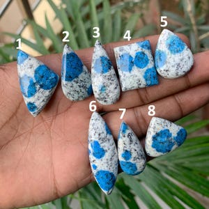 May include: A collection of eight polished K2 stone cabochons, each with a unique shape. The stones feature a speckled white base with vibrant blue orb-like patterns. The shapes include triangles, teardrops, and a square, showcasing the stone's natural beauty.