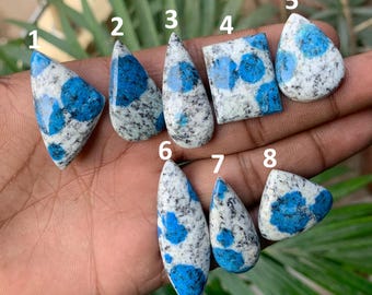 May include: A collection of eight polished K2 stone cabochons, each with a unique shape. The stones feature a speckled white base with vibrant blue orb-like patterns. The shapes include triangles, teardrops, and a square, showcasing the stone's natural beauty.